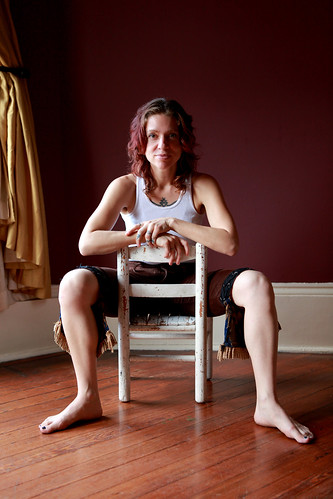 A picture of Ani Difranco sitting backwards on a chair in a red room facing hte camera. 