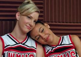 Glee characters Brittany and Santana sit in the glee club music room in their cheerleading outfits. Both smile as Santana rests her head on Brittany's shoulder.