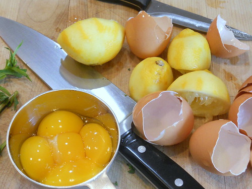 Some of the ingredients for lemon tarragon olive oil cake including 5 egg yolk in a metal bowl next to their empty brown shells. A chefs knife is on its side next to some tarragon sprigs. Several lemon halves that have had zest removed and squeezed of juice are next to the egg shells. 