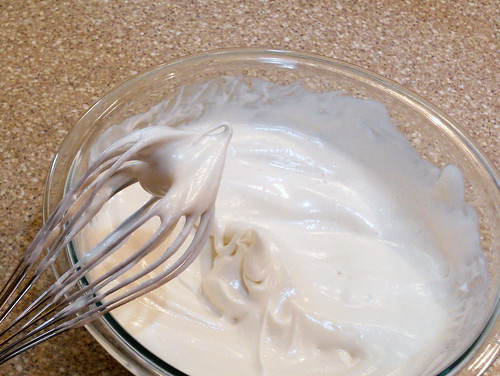 Final shot of clear mixing bowl of egg whites with whisk showing thick stiff egg whites sitting on end of whisk.