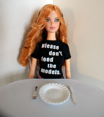 Barbie doll wearing a shirt that says "please don't feed the models" seated at a table with an empty plate in front of her.