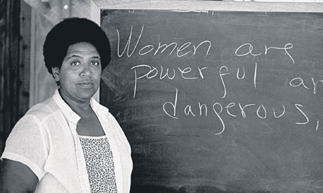 a black and white photo of Audre Lorde standing in front of a chalkboard that reads 