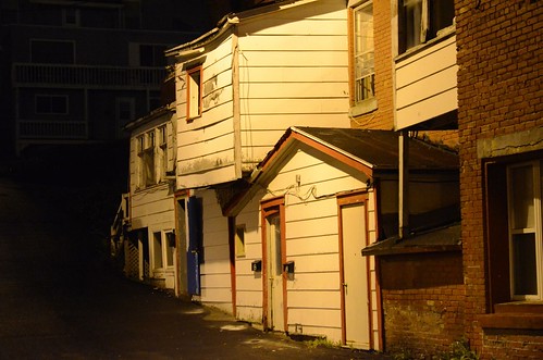 An alley in Campbellton, New Brunswick showing the rear side of houses and garage showing wear and tear, lit by street lights, and fading the dark of night.