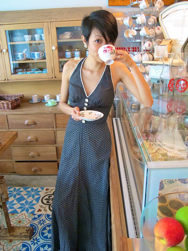 Melissa sips tea standing up, while looking grand in a black and white 1970s halter neck maxi dress with white piping. 