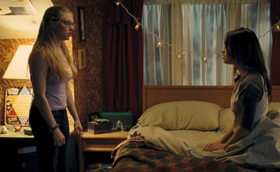 A girl with blonde hair, wearing jeans and a tank top, stands next to a bed and stares at a brunette girl, wearing a jersey, sitting on the bed. The lighting is ominous.