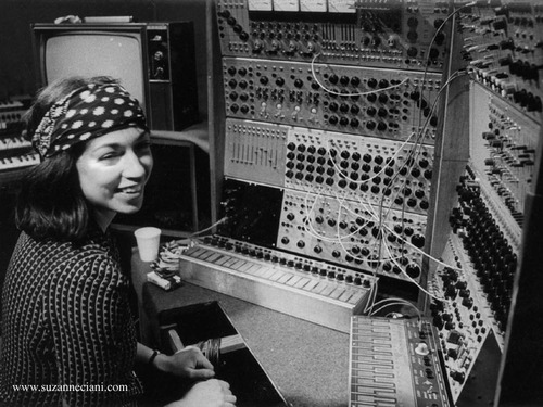 A black and white photograph of a 70s Suzanna smiling in front of a wall of knobs and wires 