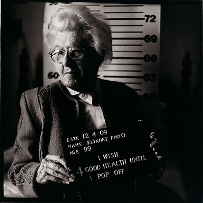 Black and white mugshot-style photo of a white woman aged 99. The placard she's holding says I wish for good health until I pop off