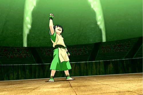 Toph standing in the middle of an empty fighting square. She triumphantly holds her fist in the air as the winner. She is wearing her typical outfit: barefoot, a green tunic with a tan tunic belted over it.