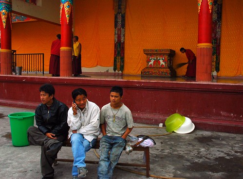 The remains of the day: three young Tibetan men sitting on a bench, taking a break, using the cell, during cleanup, monks on the stage, Tharlam Monastery courtyard and stage, long life empowerment, Boudha, Kathmandu, Nepal by Wonderlane