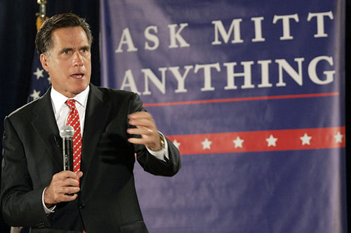 Mitt Romney in front of a sign that says ASK MITT ANYTHING. He is in a black suit with a red tie