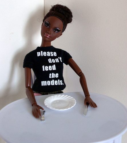 Barbie doll wearing a shirt that says "please don't feed the models" seated at a table with an empty plate in front of her.
