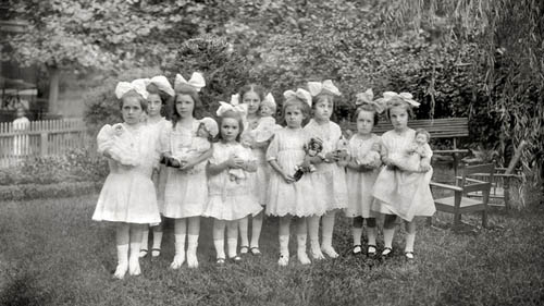 a black and white photo of a group of girls in dresses and hair bows standing outside