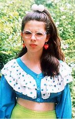 Bespectacled girl wearing red heart earrings and a floucy blue blouse.