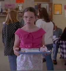 Bespectacled girl wearing a bright pink blouse and holding a lunch tray of food.