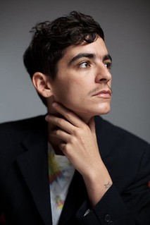 Portrait of JD Samson in front of a grey background. JD Samson is wearing a black blazer and gazing into the distance with her hand placed on her neck.