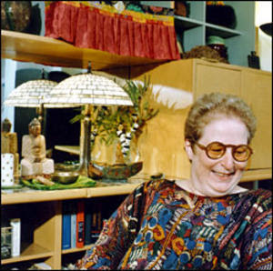 a vintage looking photo of eve sedgwick, wearing round brown glasses, and a colorful abstractly printed top, looking down to her left, and smiling