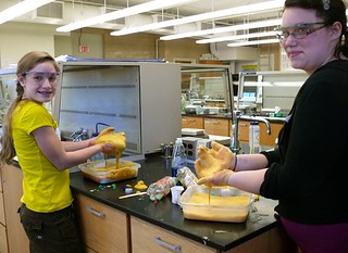Two girls work in a chemistry lab with their hands in a gooey, elephant toothpaste substance they created.