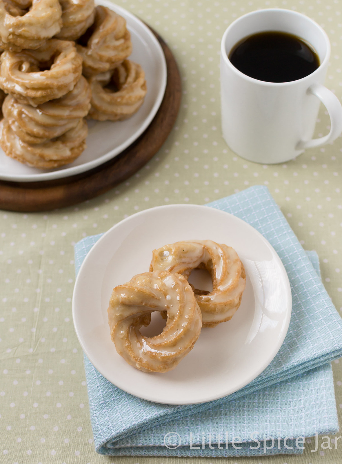 two crullers on plate with a stack in the back with coffee mug