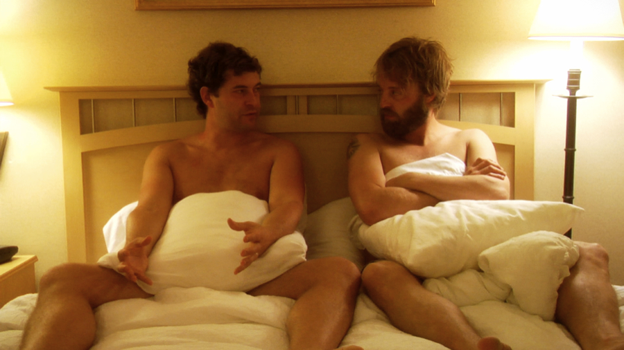Two men sit in bed together, facing each other. They are naked and pillows cover most of their bodies. Lamps are lit on either side of them.