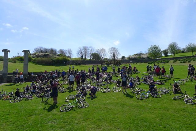 A large group of cyclists with their bikes laying all around, in a green grass field with blue sky above