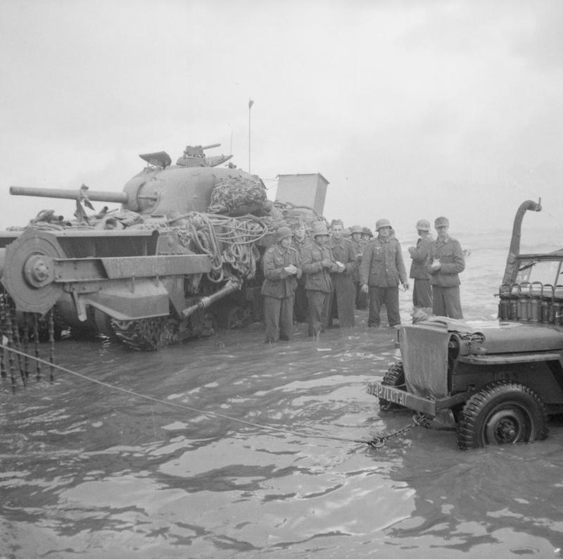A group of German prisoners standing in the water next to a disabled Sherman Crab flail tank watch as a jeep is towed from the sea, Queen beach, Sword area, 6 June 1944.