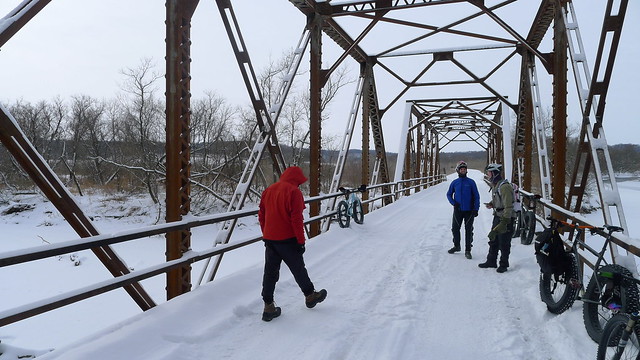 Straight away view of a snow covered bridge, with fat bikes against the sides, and cyclists walking on the deck