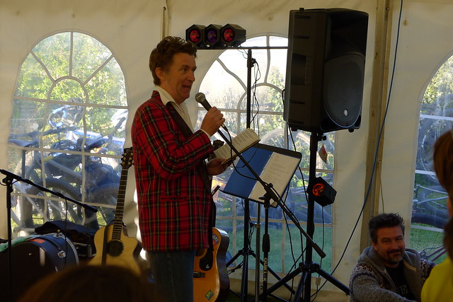 Right side view of a person in a red, plaid sportcoat, under a white event tent, speaking from microphone