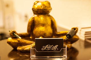 ein goldener Frisch sitzt im Schneidersitz und Meditationshaltung da und hat zwischen den Fingern Stifte, davor Visitenkarten