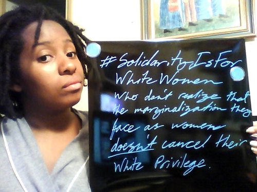 An African-American woman holding a sign that says "solidarity is for white women who don't realize that the marginalization they face as women doesn't cancel their white privilege"