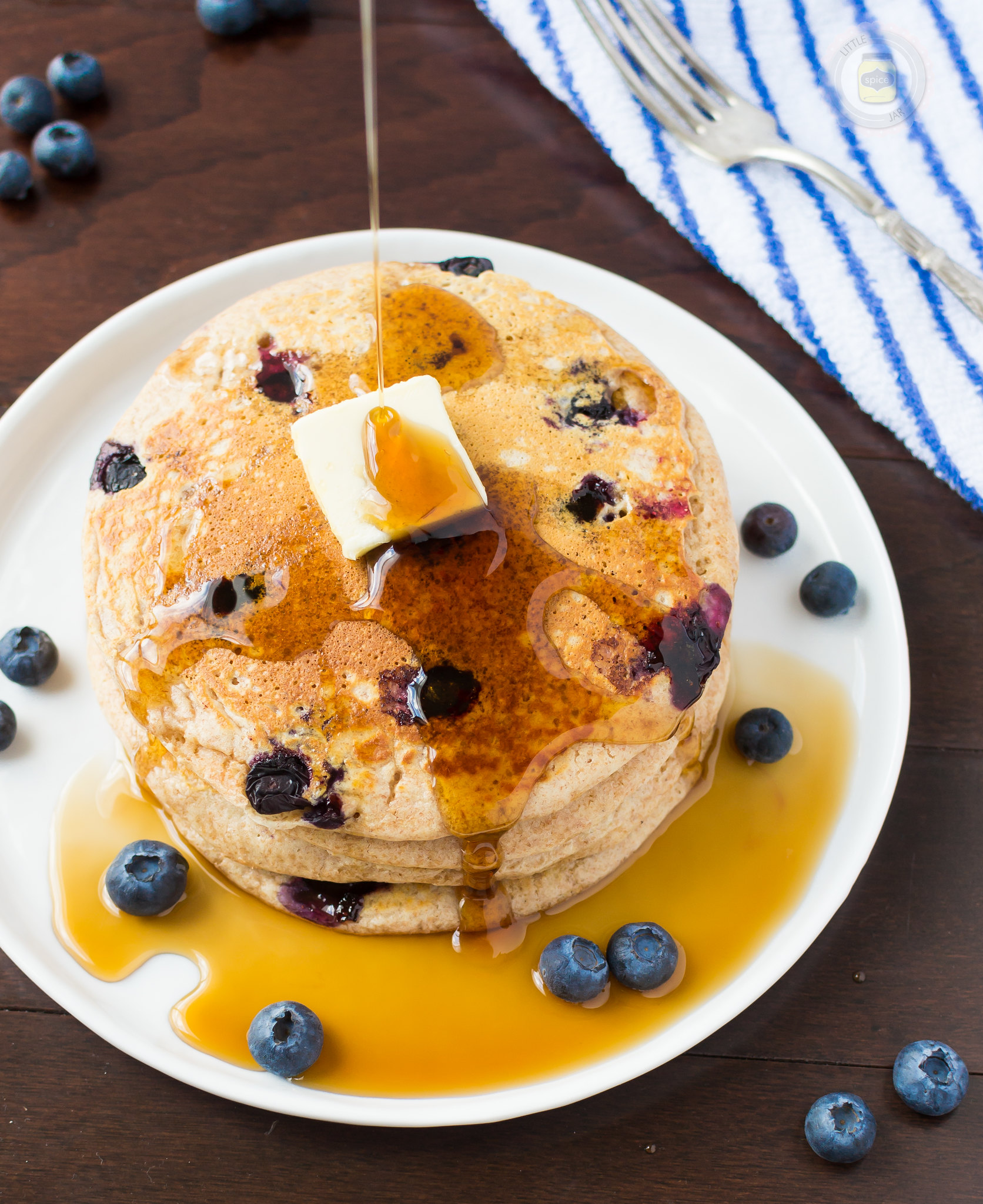 whole wheat buttermilk pancakes with blueberries on white plate with a pat of butter pouring pancake syrup on top