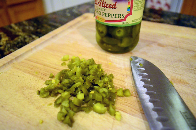 A jar of jalapeno rings on a cutting board with a small pile of diced jalapeno rings.