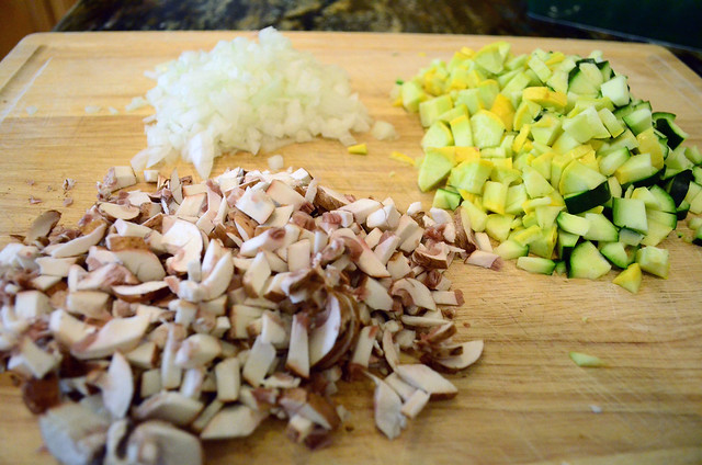 Zucchini, squash, onion, and mushrooms cut into small pieces on a cutting board.