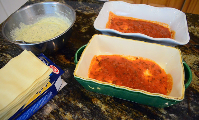 Two loaf pans with marinara sauce coating the bottom, a stack of lasagna sheet on the side and the bowl of ricotta cheese mixture.