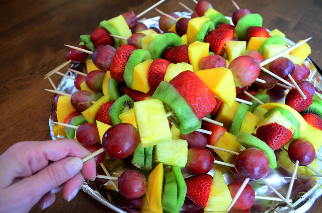 A hand holding a fresh fruit kabob over a serving plate full of fruit kabobs.