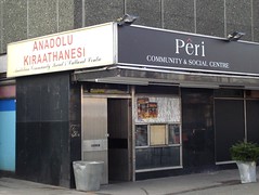 A corner unit with reinforced door and no obvious window.  The front has a sign reading “Pêri Community & Social Centre” while the side has one reading “Anadolu Kıraathanesi Anatolian Community Social & Cultural Centre”.