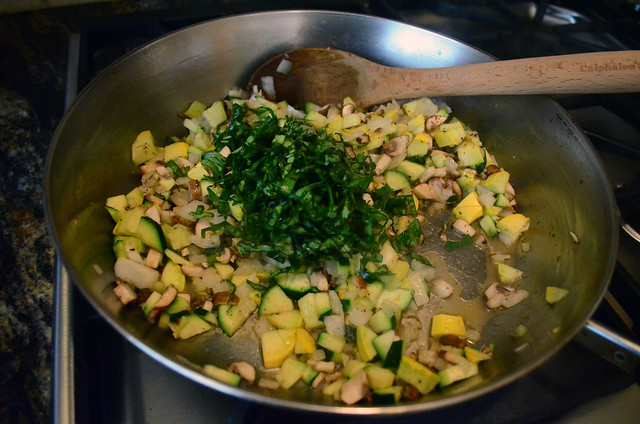 A pan with the cut veggies and chopped basil being stirred with a wooden spoon.