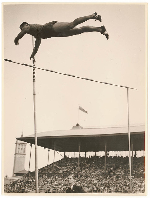 Aged black-and-white photo of a man poling over a high jump.