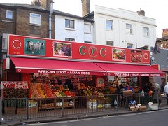 The same shop as in the first photo in this article, but stretching over two shopfronts instead of one.  The awning is longer here, and in addition reads “Vegetable — Fruit — Bakery — Grocery”.  There still seems to be a division between the two shops, at least at the front, where groceries line the wall in between.