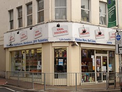 A corner shopfront painted pale cream.  Windows on the front, side, and corner display items such as wigs and hair products.