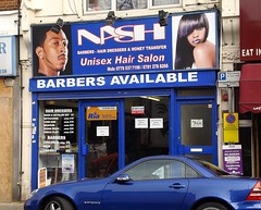 A blue terraced shopfront with a fully glazed frontage. The sign reads “Nash” and incorporates photos of two young Black people, a man and a woman, with stylish haircuts.