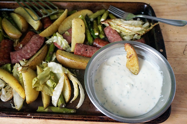 A pan of roasted vegetables and sausage, with a bowl of sauce nestled in the corner. A wedge of potato, its surface crackled with brown and gold, sinks slowly into the sauce.