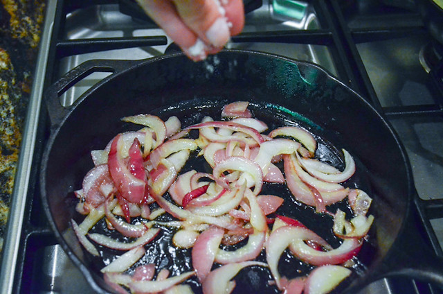 Sugar being sprinkled onto chopped onions that are cooking in a pan.