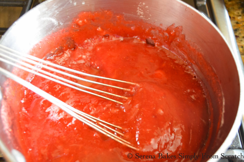 Cooked thicken strawberry filling mixture with vanilla extract being whisked in a stainless steel saucepan.