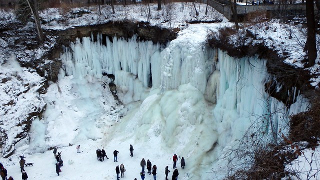 Light blue, frozen Minnehaha Falls with many people standing at the base