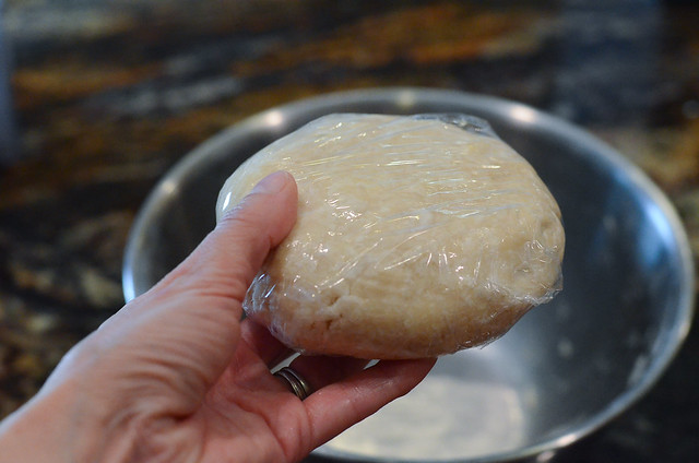 A hand holds a flattened disc of dough wrapped in plastic wrap.
