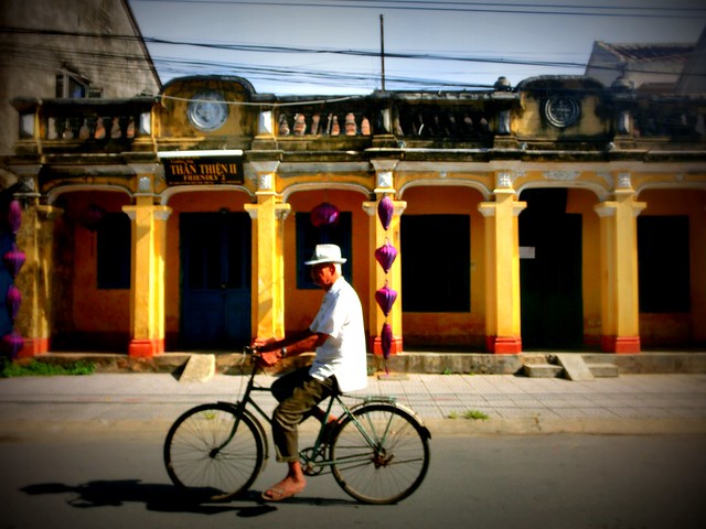 A man rides his bicycle through town with purple silk lanterns in the background