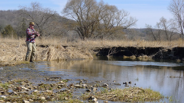 Right side view of a person, standing in a stream while fishing, with bare trees and hills in the background