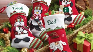 Personalized Stockings from Personal Creations with a snowman, Santa, a penguin and a dog in a basket with pine cuttings, ornaments and a small wrapped gift with a green bow