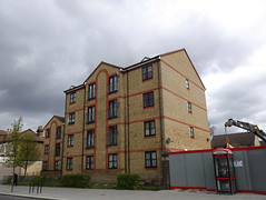 A four-storey building in light brown brick with decorative brick detailing in a darker orange shade.  There are several windows on each level, but no obvious entrance on this side.  To the right is a grey hoarding with a phone box in front and a crane behind.