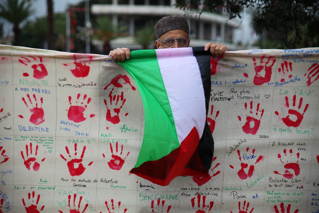 一位行動者在遊行中揮舞巴勒斯坦國旗，一個個的血手印象徵在以色列攻擊中遭殺害的受難者。（攝影：陳逸婷）